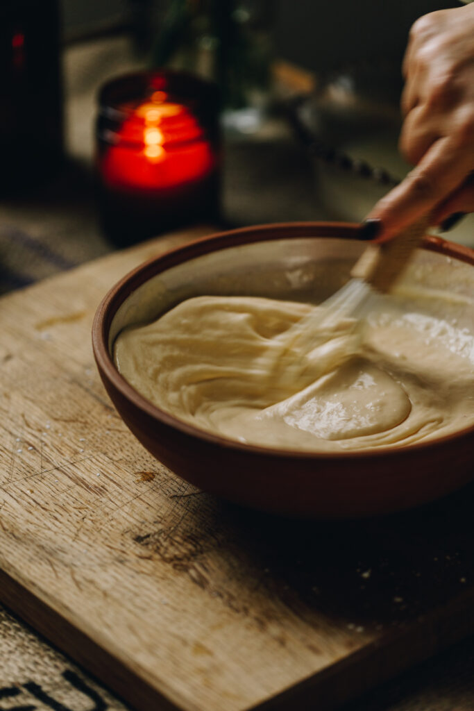 A clay bowl sits on top of a wooden board with an amber candle in the background. In the bowl is an action shot of the Lemon Meringue Pie custard being stirred. 