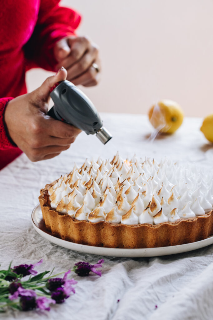 On a tablecloth sits a freshly piped lemon meringue pie that is being torched with a chef's torch. Lavender flowers are in the forefront and lemons are in the back. 