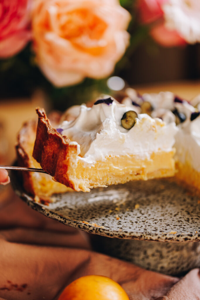 A piece of homemade lemon meringue Pie is being held up with a cake server. It is being lifted off a ceramic cakes stand with orange flowers in the background. 