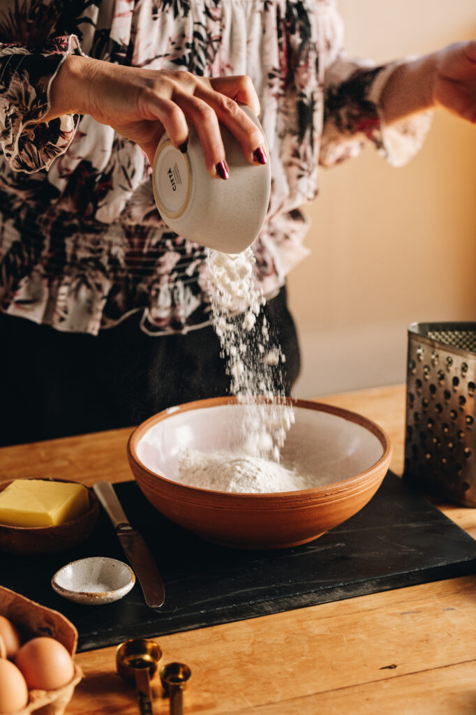 A clay bow sits on a black board. An action shot shows flour being tipped from a small bowl in to the clay bowl. 