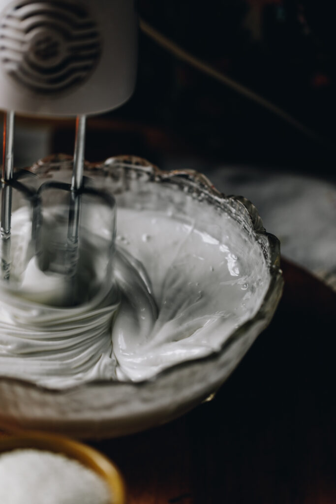 A glass bowl is filled with a meringue that is being whipped by a hand beater for the How to make Italian Meringue recipe. It is glossy and the shot is moody.