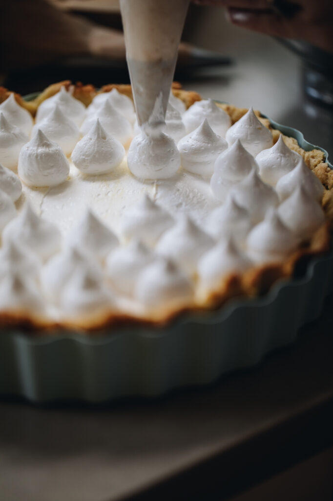 The Italian Meringue is being piped on to a pie. The shot is close and shows the glossy meringue piping perfect peaks on to the pie.