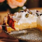 A close up of slice of homemade lemon meringue pie is suspended in the air as it is lifted of a grey ceramic cake plate.
