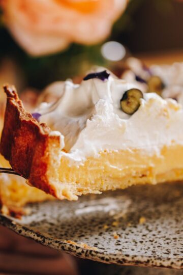 A close up of slice of homemade lemon meringue pie is suspended in the air as it is lifted of a grey ceramic cake plate.