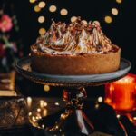 A decorated smores no bake chocolate cheesecake is on a blue plate on top of a red glass cake stand. Glowing fairy lights are in the background.