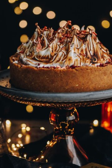 A decorated smores no bake chocolate cheesecake is on a blue plate on top of a red glass cake stand. Glowing fairy lights are in the background.