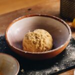 A ball of sweet lemon pastry sits in a ceramic bowl on top of a black board that is sprinkled with flour.