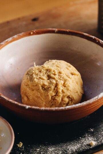 A ball of sweet lemon pastry sits in a ceramic bowl on top of a black board that is sprinkled with flour.