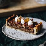 A slice of pean pie with dark chocolate sits on a grey speckled ceramic plate. It is decorated with piped yogurt, small apples and gold leaf.
