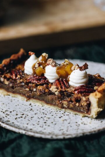 A slice of pean pie with dark chocolate sits on a grey speckled ceramic plate. It is decorated with piped yogurt, small apples and gold leaf.