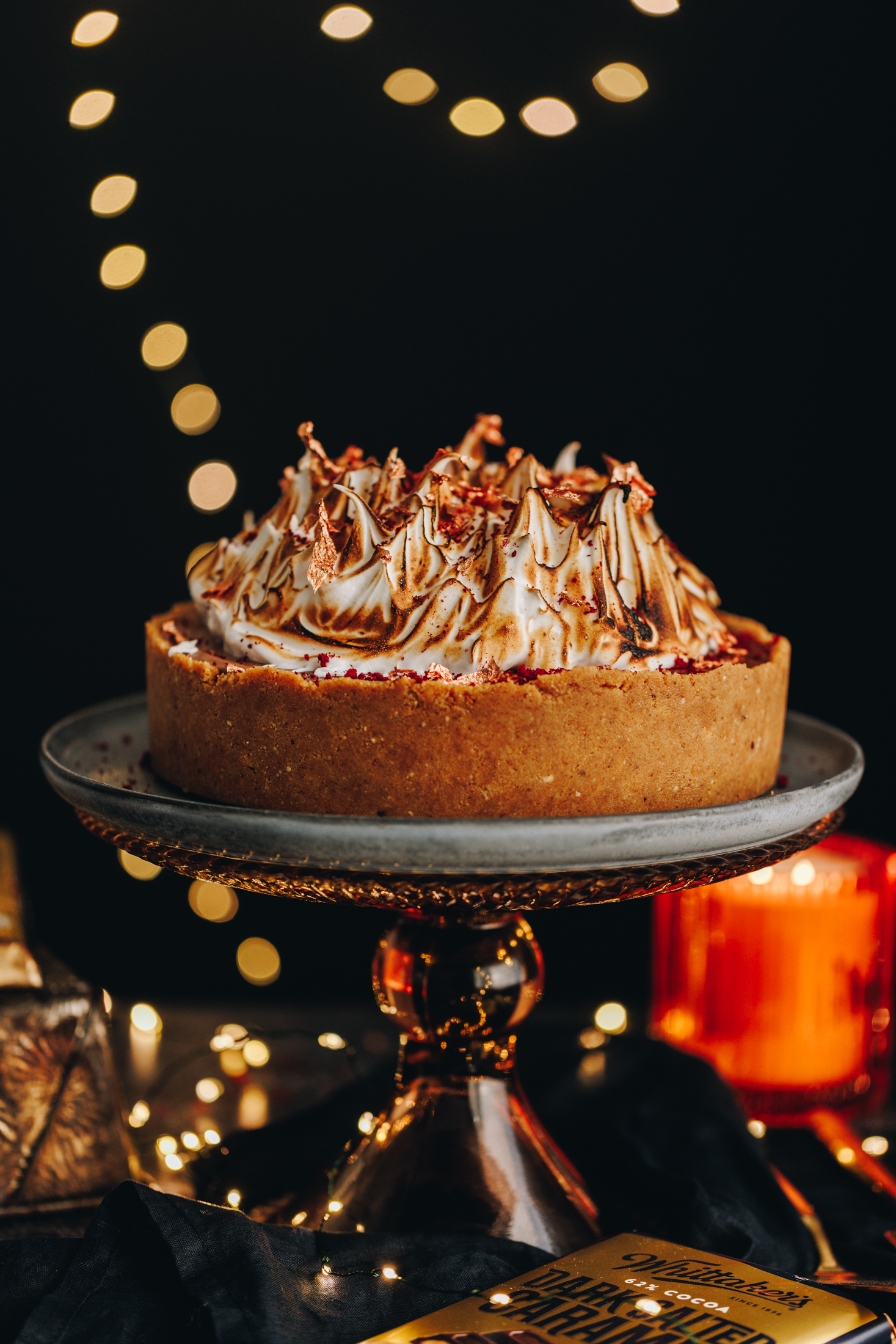 A S'mores No-bake Chocolate Cheesecake is on a glass cake stand with a blue plate on it. It is decorated with meringue and lights glow in the background.