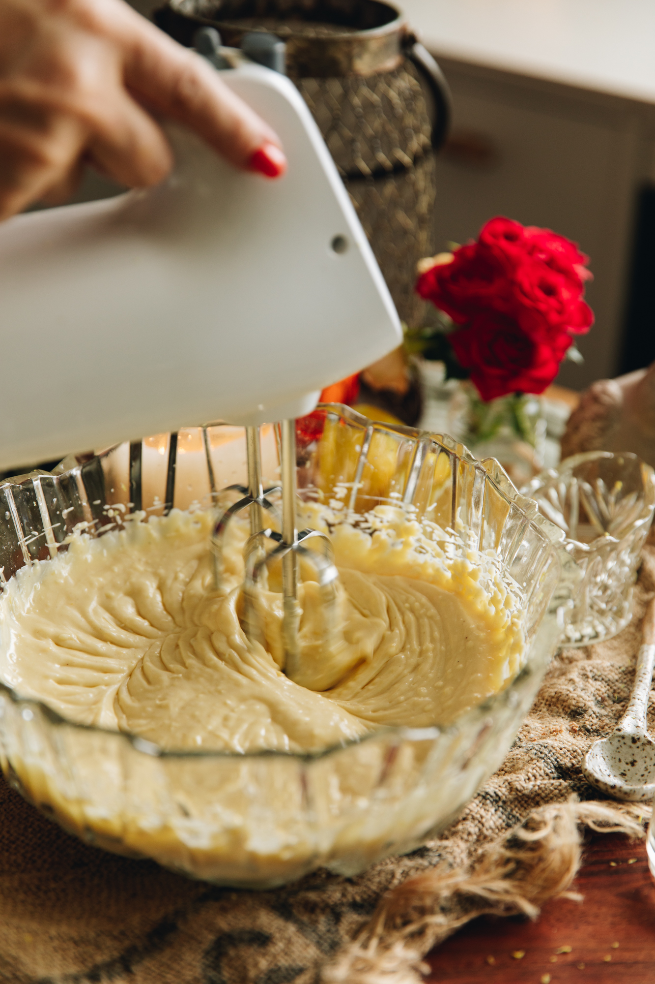 A glass bowl on a wooden table is whipping cream cheese with a white hand mixer. Bright flowers are in the background.