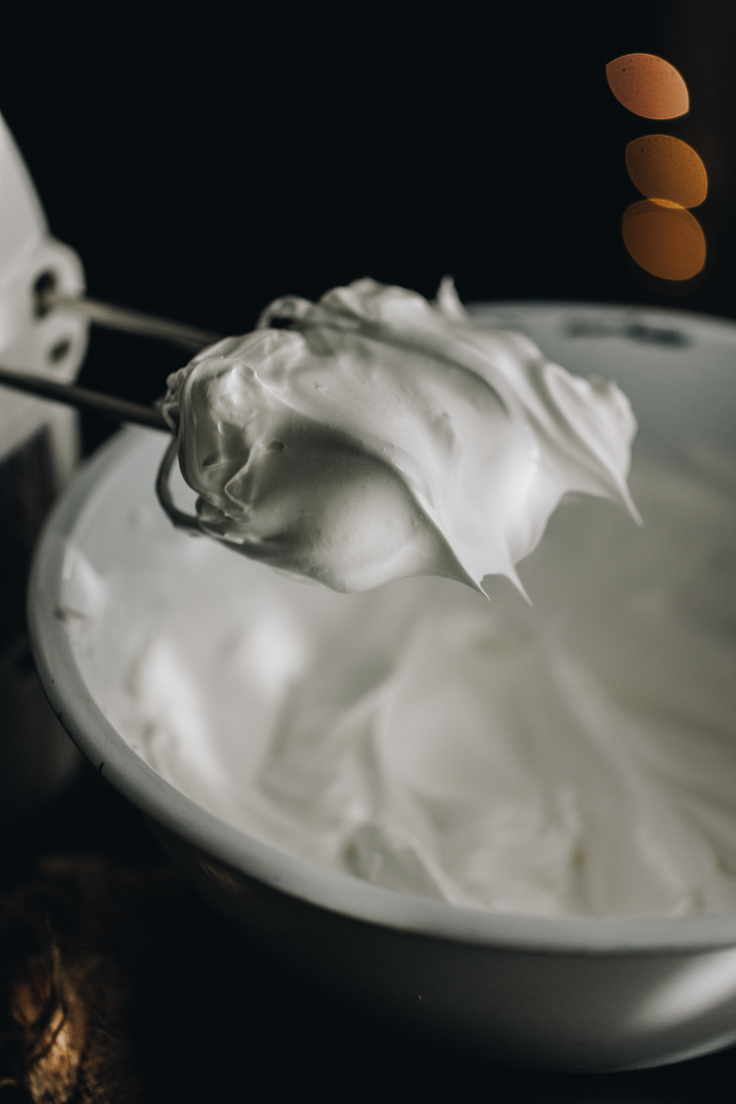A meringue is in a white bowl and a hand mixer holds stiff peaks of Italian meringue at the tip of the beaters.