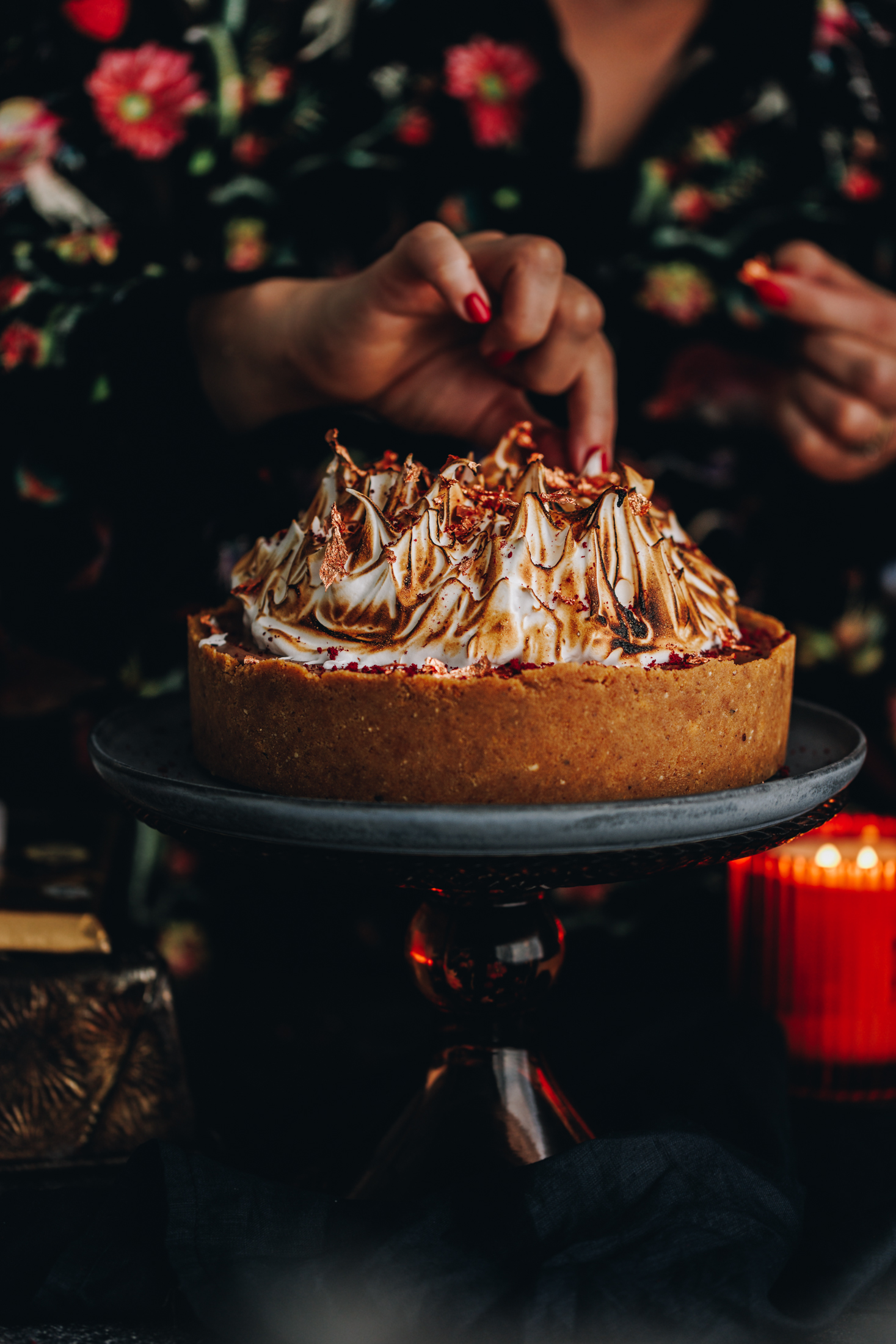 Naomi is decorating the S'mores No-bake Chocolate Cheesecake by adding gold leaf on top of the torched meringue. The cake is on top of a glass cake stand.