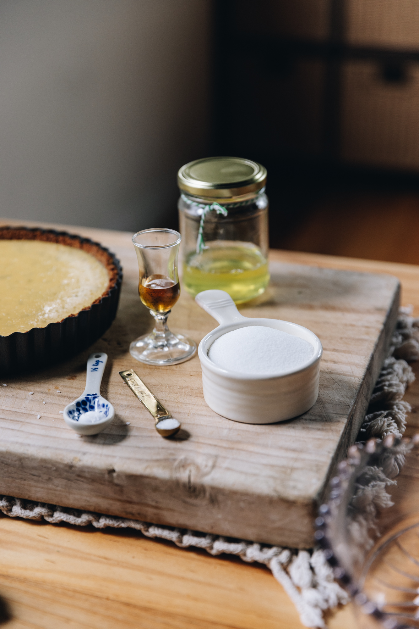 On a wooden board sits vintage cups and spoons that are holding the ingredients for the Italian meringue. A baked lime pie is on the side of the board. It sits on a cream weaved mat, on top of a wooden table.