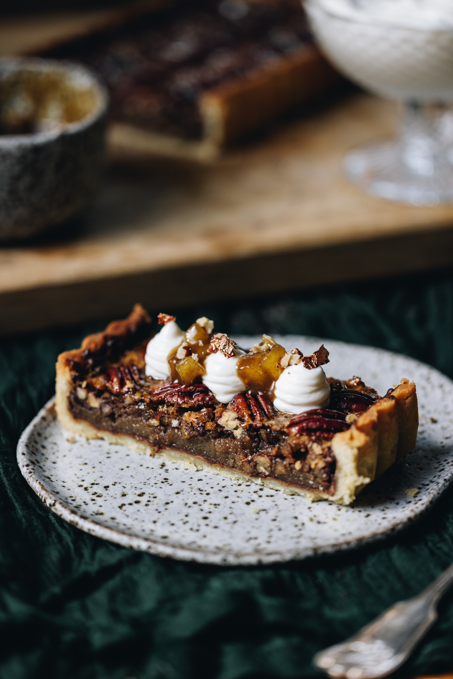 On a ceramic plate sits a slice of pecan pie with dark chocolate. It has been decorated with piped yogurt, caramelised apple and some gold leaf. The plate is on a green cloth.