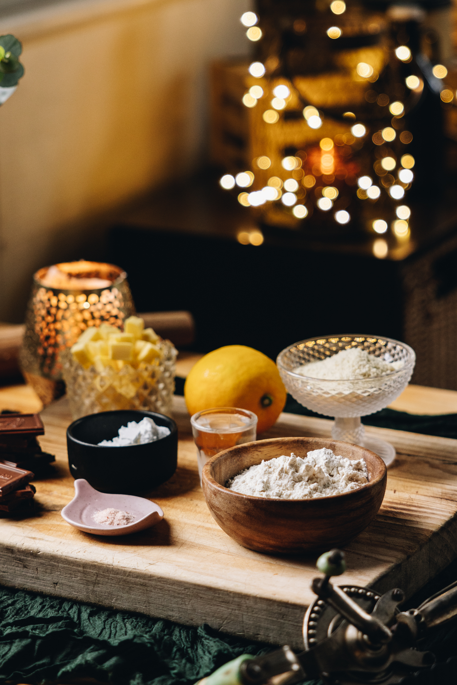 On a wooden board sits vintage bowls and vessels holding the pie ingredients. There is flour, butter, a lemon and chocolate in view. A golden candle burns in the background and golden fairy lights are also seen.