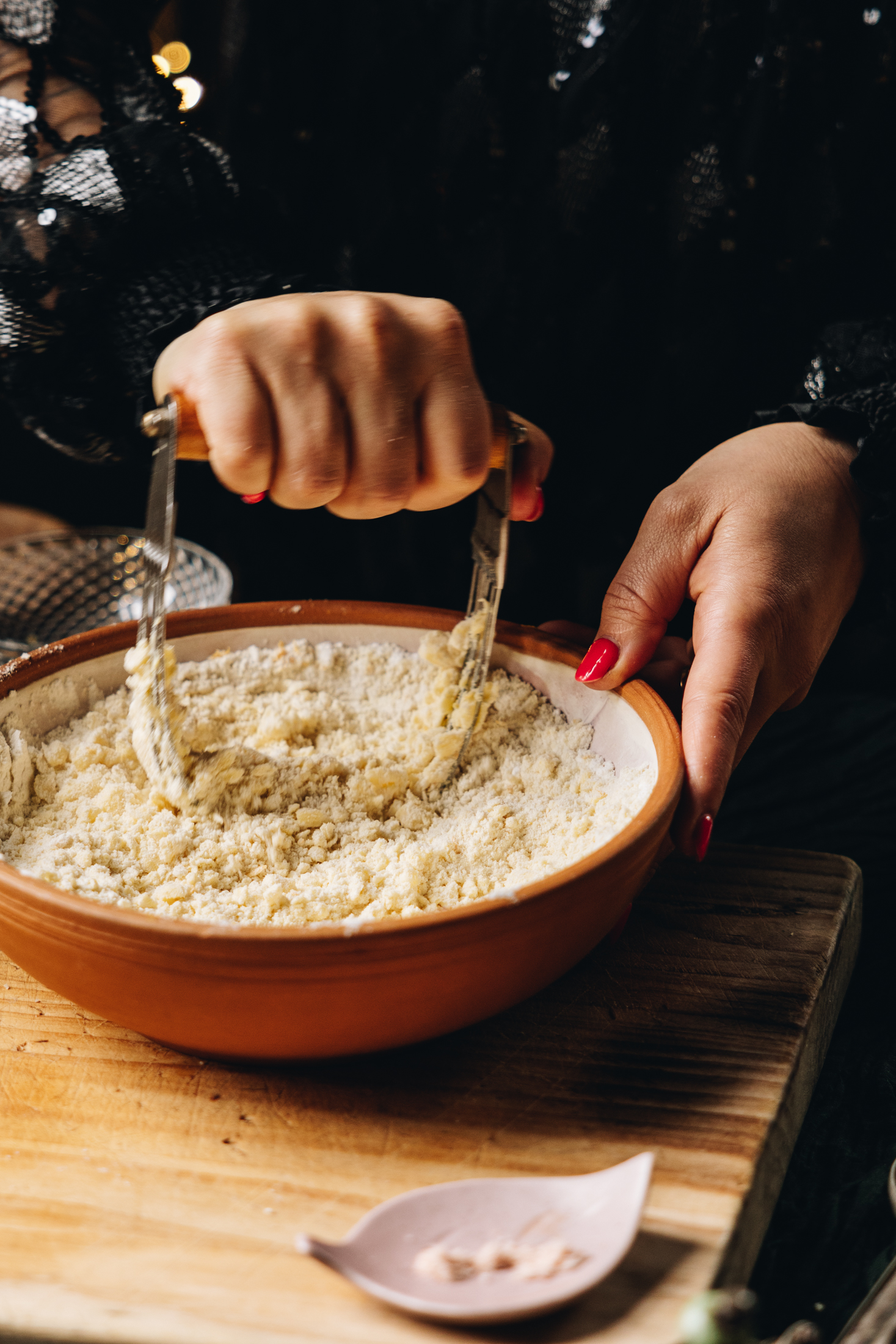 A ceramic bowl sits on a wooden bench with a leaf shape small plate with salt next to it. Naomi is holding a pastry cutter and is pressing butter in to a flour mixture.