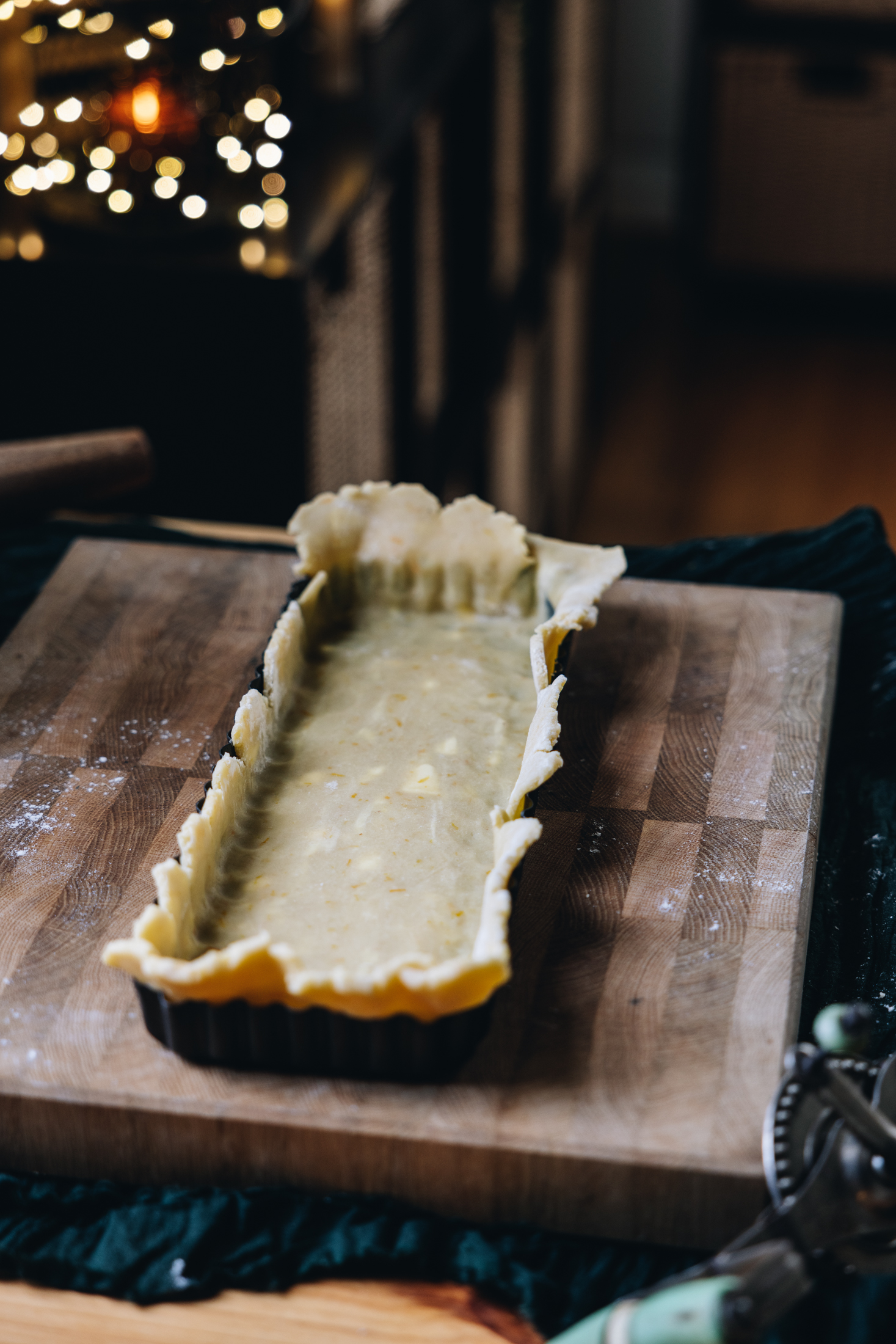 On a wooden board sits a long rectangle black quiche tin that is lined with pastry with a large overhang. The board is on a table with a green cloth underneath it. Fairy lights are seen in the background.