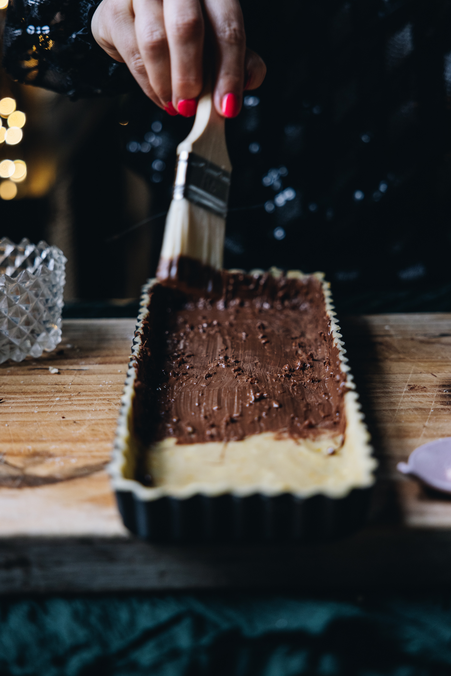 On a wooden board sits a long rectangle black quiche tin that is lined with pastry that has been trimmed and is now being brushed with chocolate with a pastry brush. Half of the pastry is covered with chocolate and the rest is bare. The board is on a table with a green cloth underneath it. Fairy lights are seen in the background.