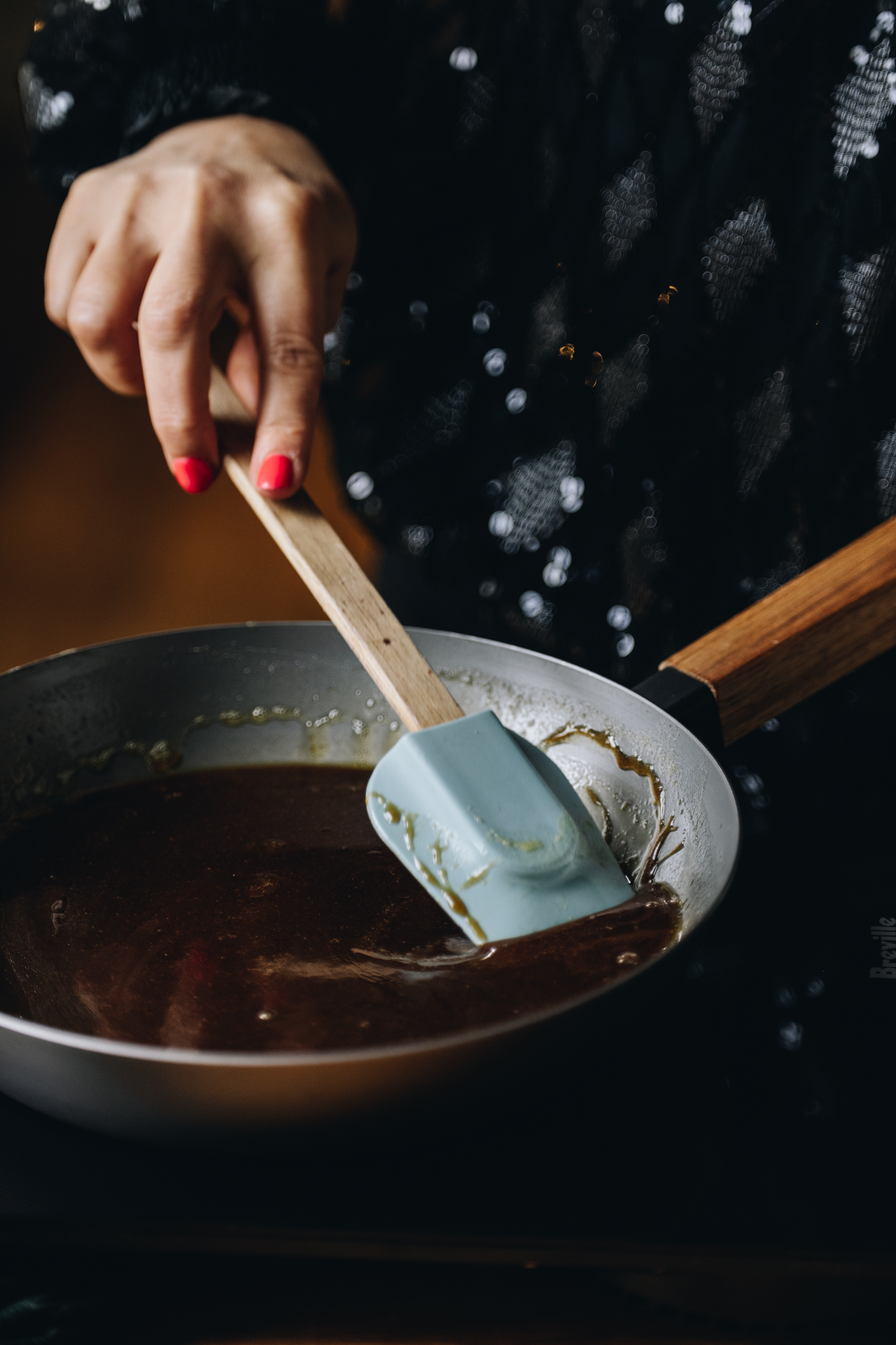 A grey cast iron pan sits on a black portable stove. In the pan is maple syrup caramel that Naomi is stirring with a blue spatula with a wooden handle.