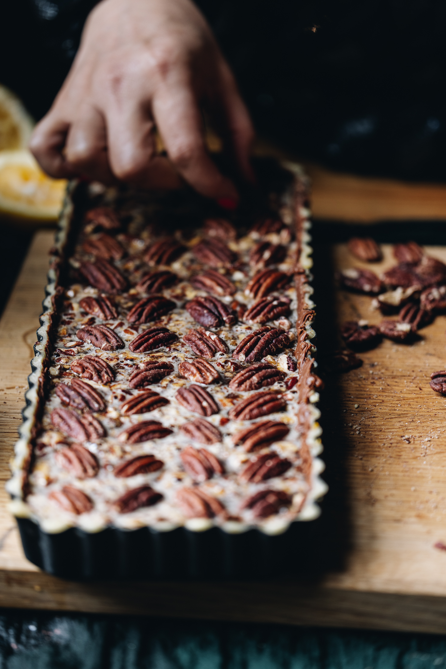 On a wooden board sits a long rectangle black quiche tin that is lined with pastry that has been trimmed and is now having whole pecan nuts arranged the top in a herringbone pattern. Naomi is placing a nut at the end of the pie, finishing off the deign.