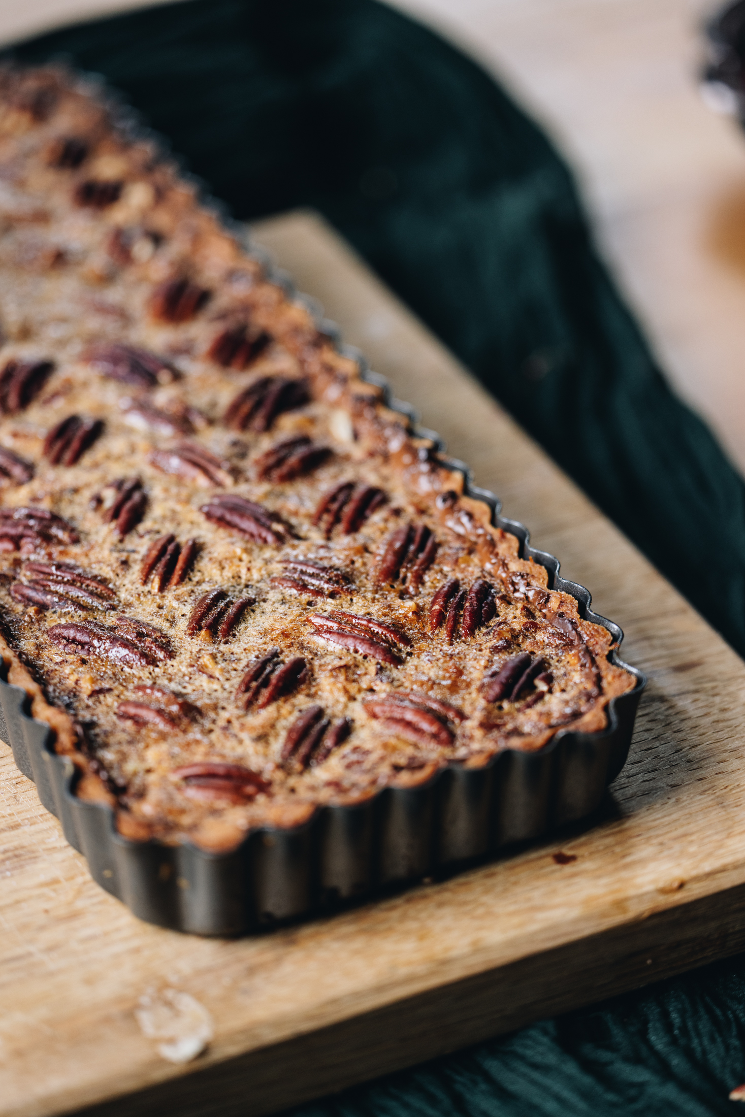 On a wooden board sits a long rectangle black quiche tin that has a freshly baked pecan pie with dark chocolate on it. The pecan nuts on top have been arranged in a herringbone pattern.