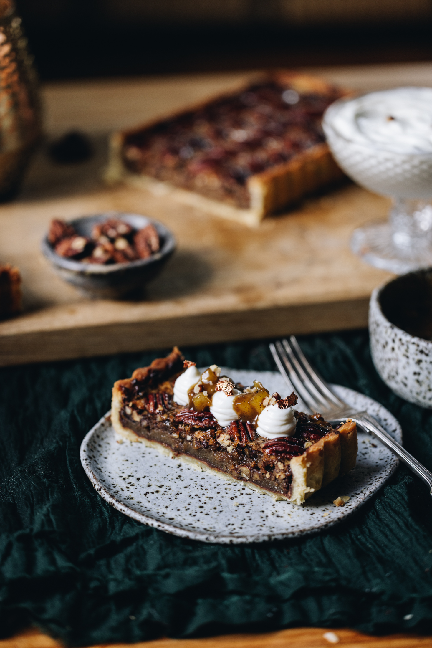 On a ceramic plate sits a slice of pecan pie with dark chocolate. It has been decorated with piped yogurt, caramelised apple and some gold leaf. The plate is on a green cloth and a whole cut pecan pie is sitting in the background with a cream in a glass jar and a little bowl of pecans.
