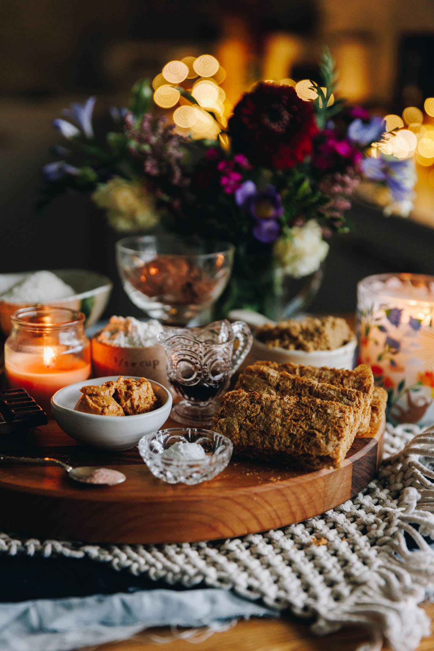 The Weetbix Slice ingredients displayed on a wooden board and multiple cloths with a candle and flowers in the background. There is salt, peanut butter, flour, vanilla and brown sugar in view.