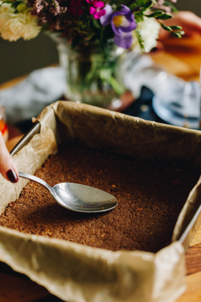 The Weetbix Slice base has been pressed in to a baking tray lined with baking paper and a spoon is smoothing out the base to flatten it. 