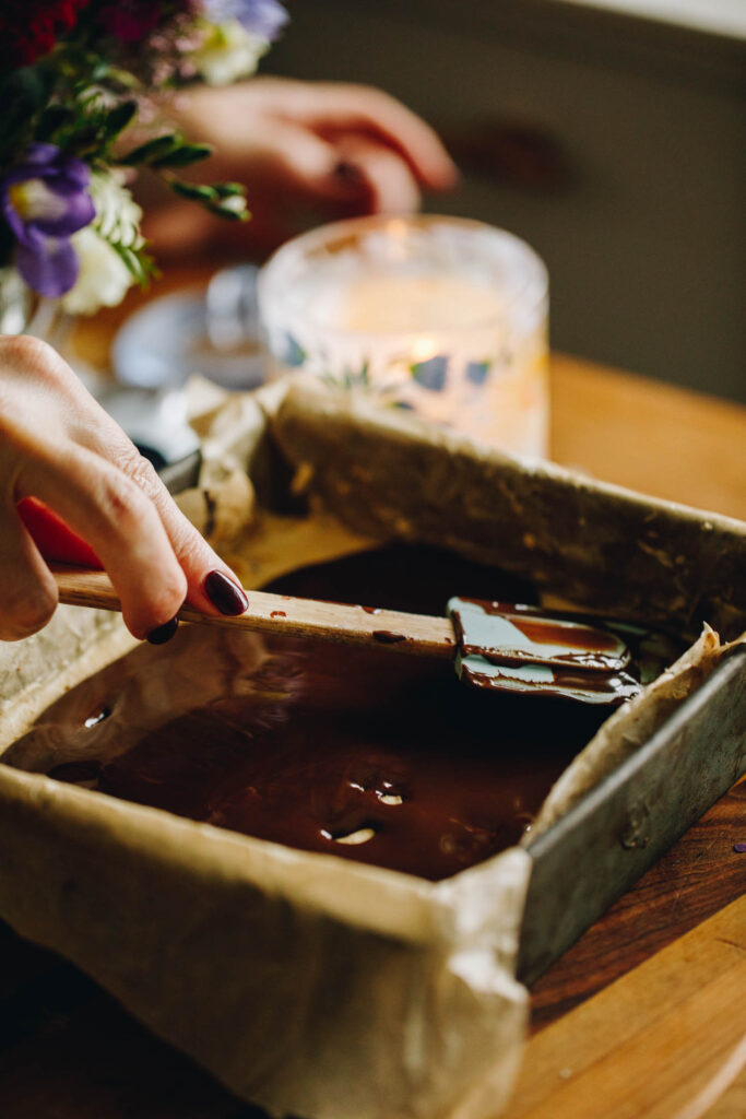 The chocolate topping is being spread on the peanut buttercream part of the Peanut Butter Weetbix Slice and is being smoothed out with a wooden spatula.
