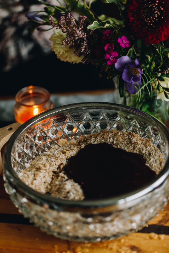 Sitting on a wooden board is a glass bowl with a silver trim. Inside is a coconut mixture with a wet chocolate mixture sitting in the centre. 