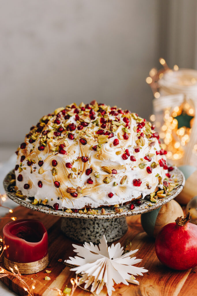 The Cheat's Bombe Alaska is plated on a ceramic cake stand surrounded by pomegranate's, a candle and a Christmas decoration. The bombe Alaska is covered in meringue and is studded with pomegranate seeds and chopped pistachios.