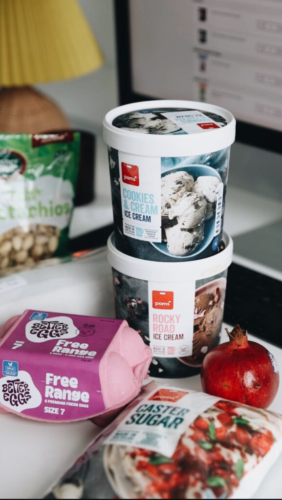 Two ice cream tubs are stacked on top of each other with a 6 pack of free range eggs, a packet of caster sugar and a whole pomegranate on a white bench. Pistachios are blurred in the background.