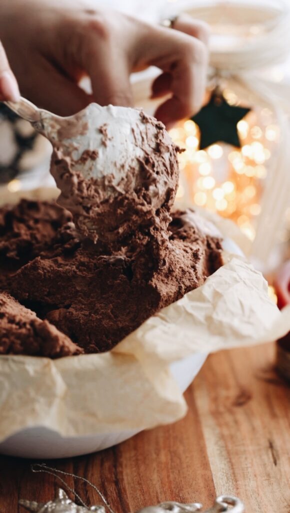 A white bowl is on a wooden board. Chocolate ice cream for the Bombe Alaska is being pressed in to the bowl with a large silver spoon. I star and twinkly lights are in the background.