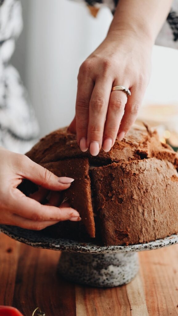 Two hands are draping pieces of cut chocolate sponge on top of the Cheat's Ice Cream Bombe Alaska. It is placed on a ceramic cake stand, on top of a wooden board.