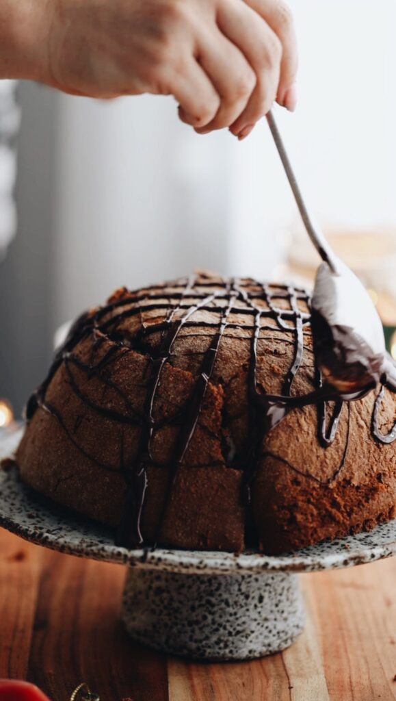 A spoon is drizzling dark chocolate all over the Cheat's Ice Cream Bombe Alaska. It is placed on a ceramic cake stand, on top of a wooden board.