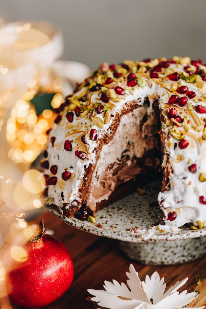 The Cheat's Ice Cream Bombe Alaska is placed on a ceramic cake stand, on top of a wooden board and a piece has been cut out of it, revelling the melting two layered ice cream inside it.