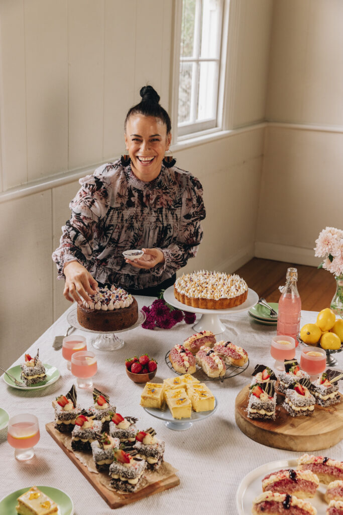 Naomi Toilalo is at the table laden with baking. There is lamingtons, lemon meringue pie, custard slice and cream buns. She is decorating a cake in front of her and smiling.