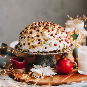 The Cheat's Bombe Alaska is plated on a ceramic cake stand surrounded by pomegranate's, a candle and a Christmas decoration. The bombe Alaska is covered in meringue and is studded with pomegranate seeds and chopped pistachios.
