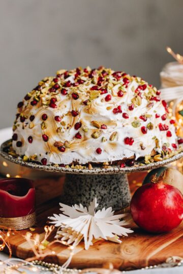 The Cheat's Bombe Alaska is plated on a ceramic cake stand surrounded by pomegranate's, a candle and a Christmas decoration. The bombe Alaska is covered in meringue and is studded with pomegranate seeds and chopped pistachios.