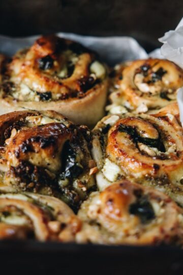 Baked cheese and pesto scrolls sit in a black tray, white baking paper peeps out from the tray.