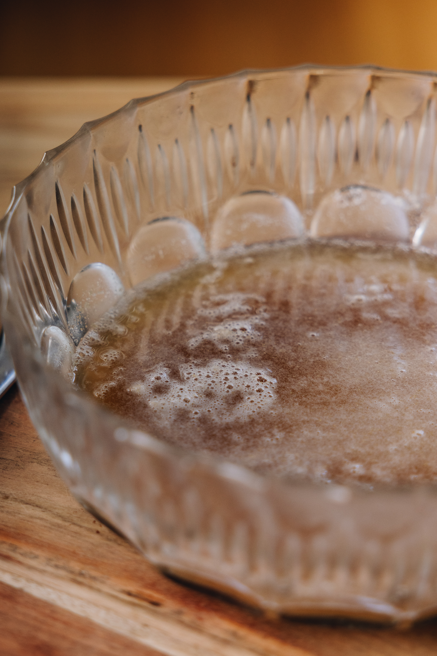 A bowl of activated yeast sits on a wooden table. The yeast is lightly foamy and rising to the top of the water.