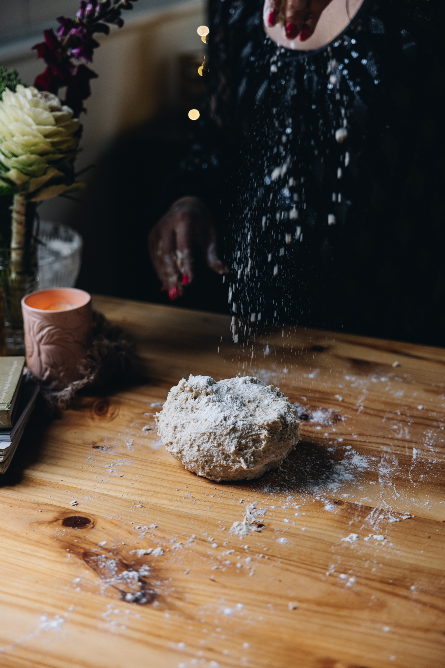 A sticky dough is on a wooden table, it is in a rough ball and hands are sprinkling flour from above on top of the dough. 