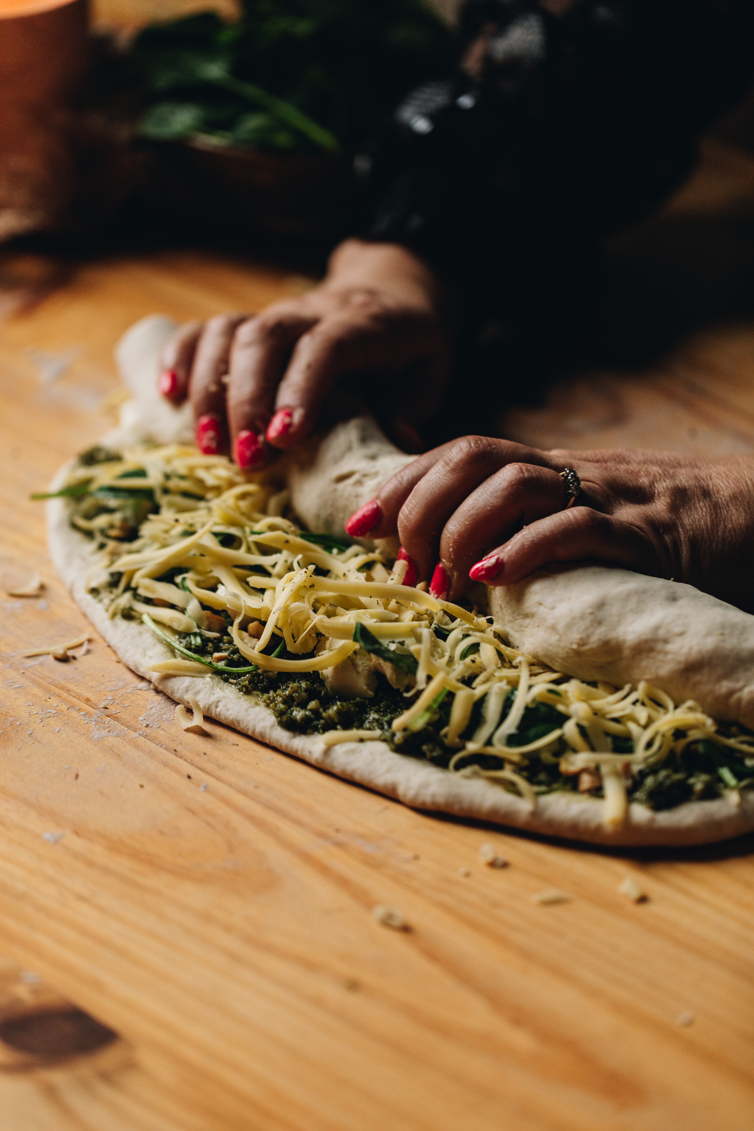 The rolled out cheese and pesto scroll dough has been topped with pesto and cheese. Two hands are rolling up the dough with some of the filling still visible. 