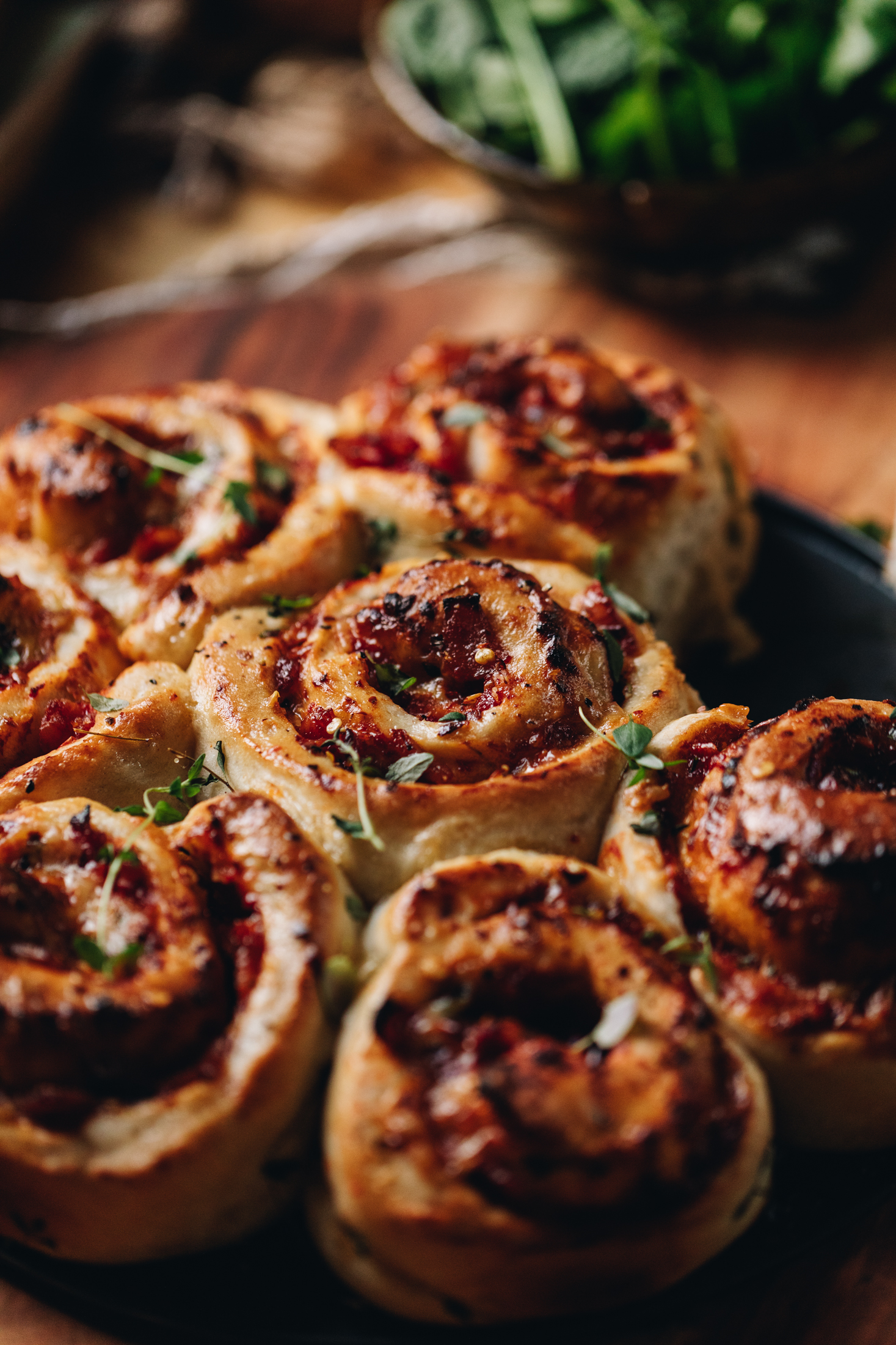 Close up of Cheese and Tomato Scrolls that are freshly baked. They have tomato and fresh thyme on them and are sitting on a wooden table with fresh greens blurred in the background.