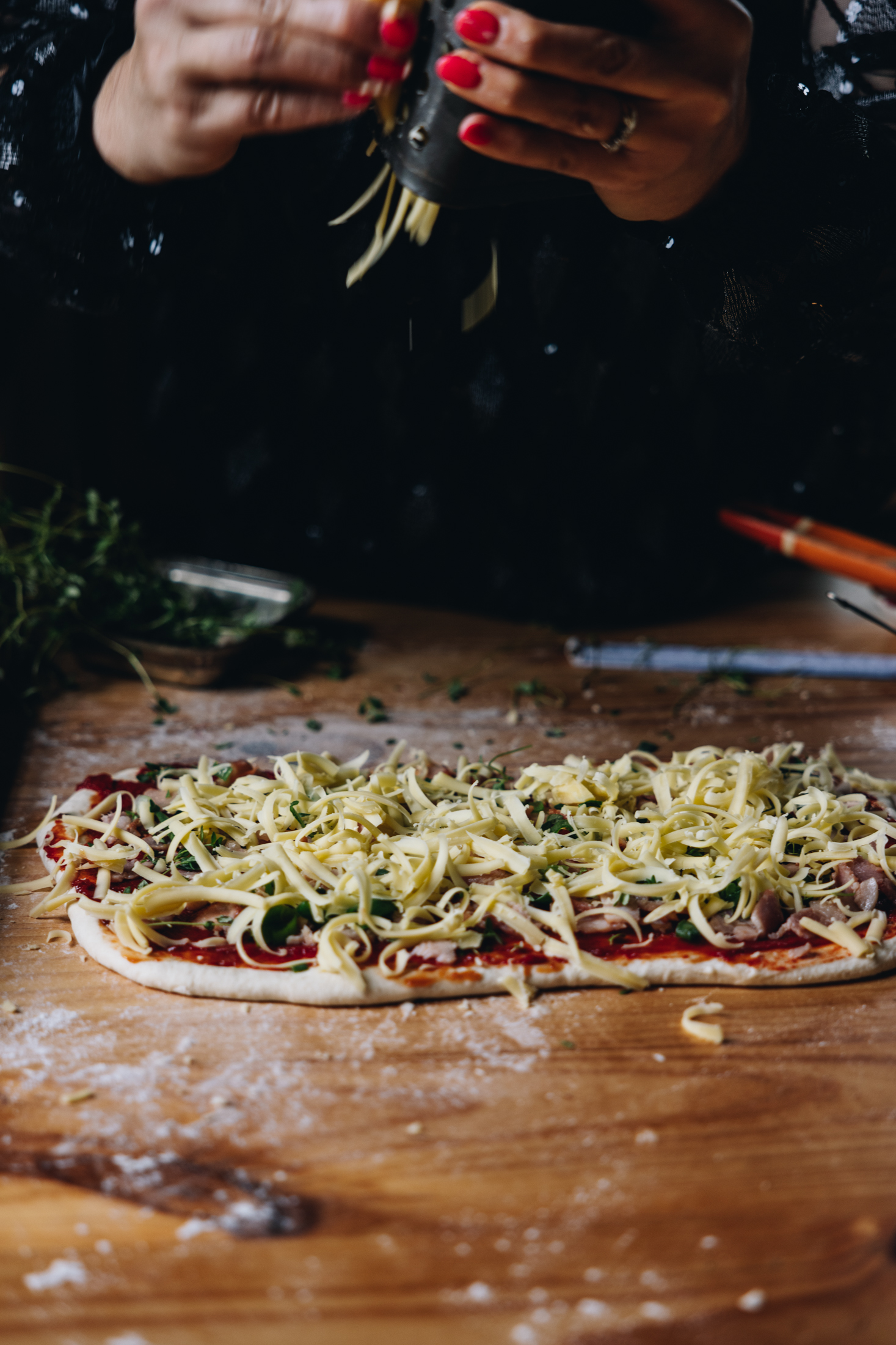 The rolled out cheese and tomato scroll dough has been topped with tomato sauce, bacon and is having cheese grated on to it. 