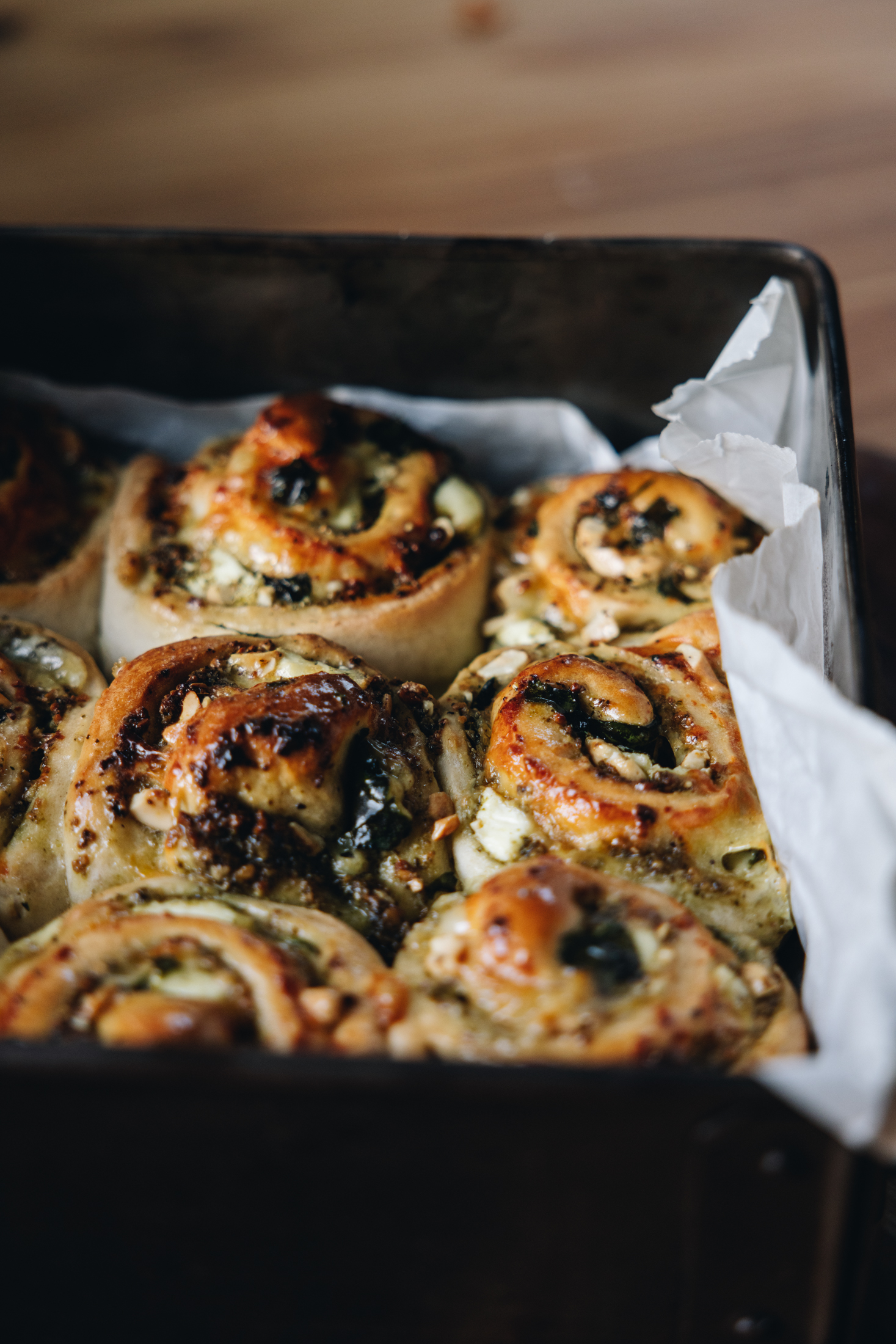 Baked cheese and pesto scrolls sit in a black tray, white baking paper peeps out from the tray.