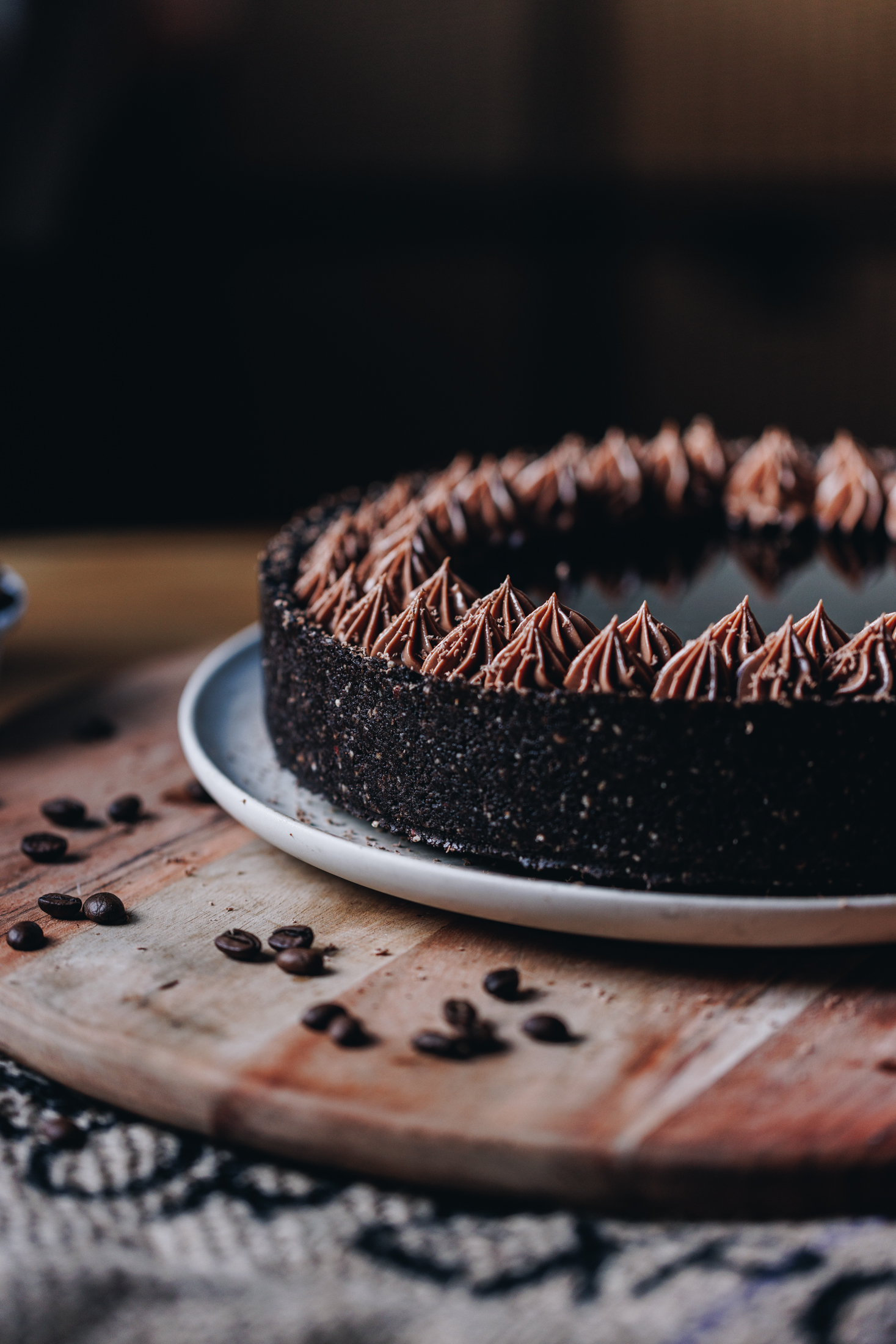 A whole chilled mocha cheesecake is sitting on a pink plate, that is on a wooden board with coffee beans scattered over it. The cheesecake has a dark crust and chocolate piped cream on top, in the centre is a coffee syrup. 
