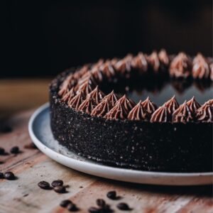 A whole chilled mocha cheesecake is sitting on a pink plate, that is on a wooden board with coffee beans scattered over it. The cheesecake has a dark crust and chocolate piped cream on top, in the centre is a coffee syrup.